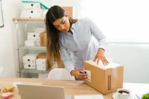 mulher colando etiqueta de envio em um pacote sobre uma mesa. Ao fundo, caixas brancas e cruas de papelão. Na mesa, uma xícara de café, um bloco de notas e um computador desfocado.